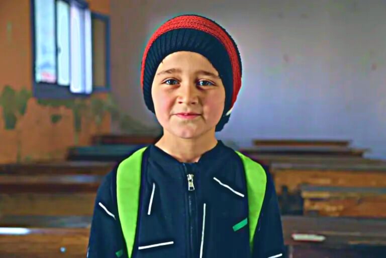 Child in classroom with backpack and beanie.