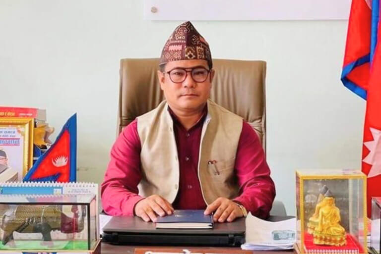 Man in traditional Nepali attire sitting at desk with flags.