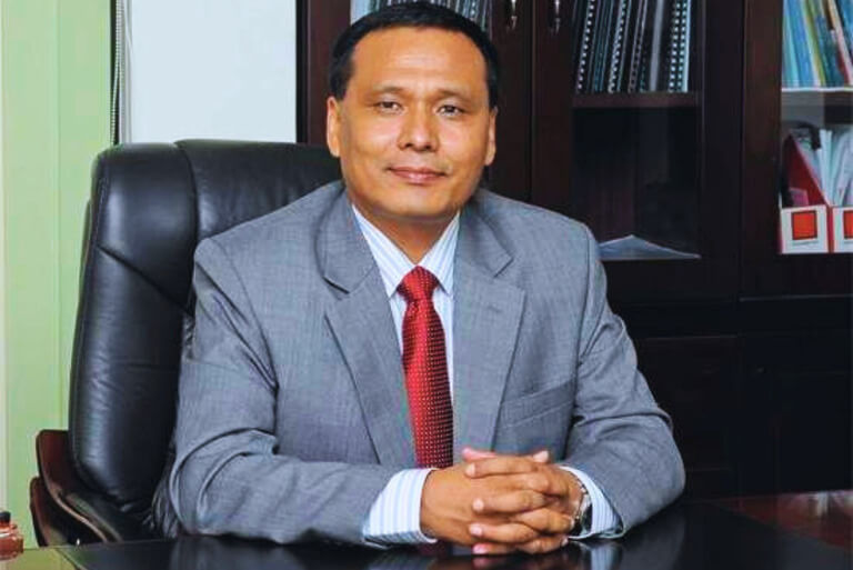 Man in a gray suit with red tie, sitting at a desk in office.