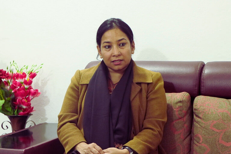 Woman in brown coat on a sofa, next to vibrant flowers.