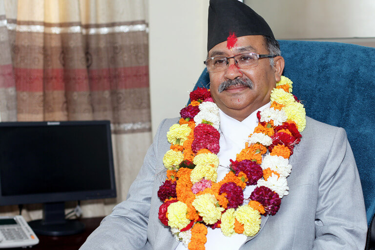 Man in traditional attire wearing a floral garland, seated indoors.
