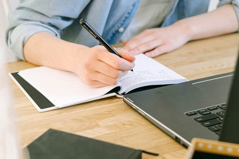 Person writing notes next to a laptop on a wooden desk.