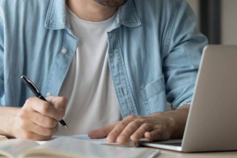 Person writing notes beside a laptop.