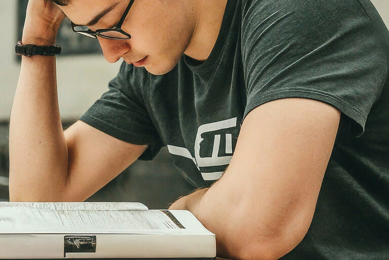 Man reading a book intently at a desk, wearing glasses.